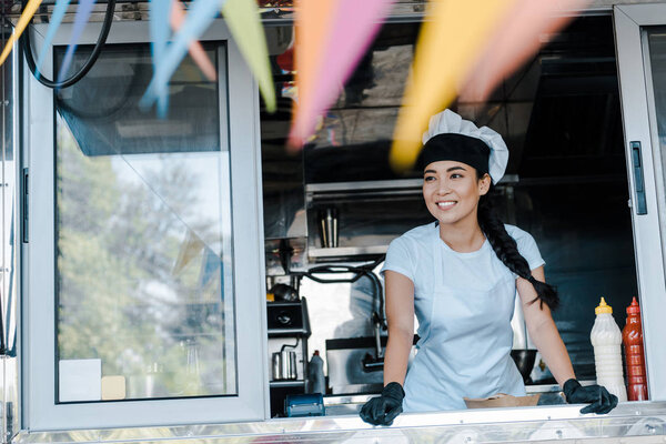 selective focus of happy asian woman in hat and chef uniform smiling in food truck 