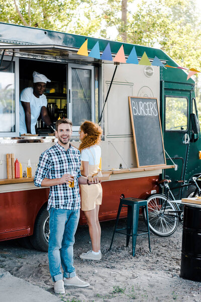 happy man holding bottle with beer near woman and african american man in food truck 