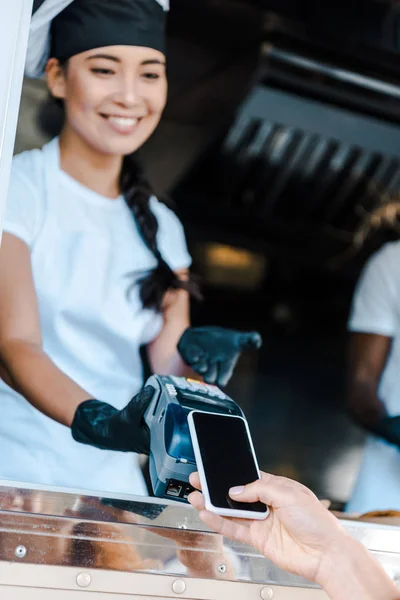 フードトラックで幸せなアジアの女の子の近くにスマートフォンで支払う女性のトリミングビュー — ストック写真