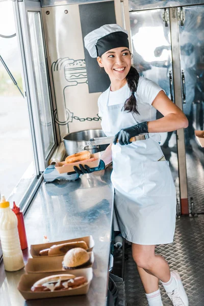 Szczęśliwy Asian Girl Holding Karton Podczas Umieszczania Hot Dog — Zdjęcie stockowe