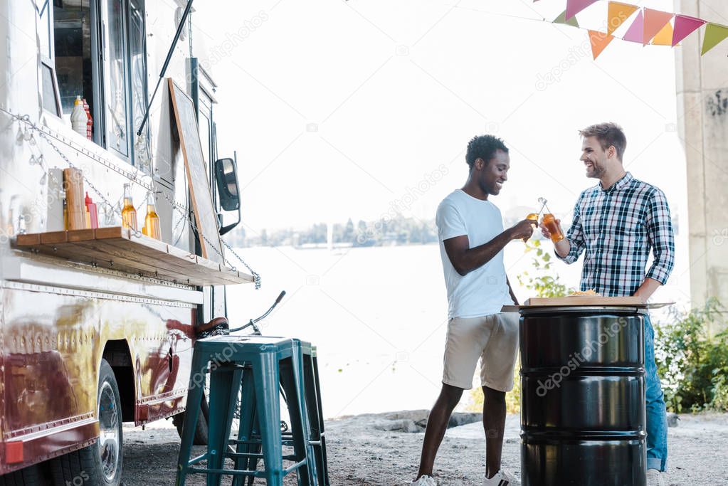 positive multicultural men clinking bottles of beer near food truck outside 