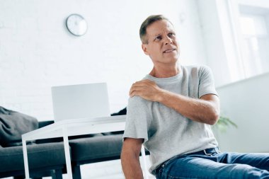 handsome man in t-shirt feeling pain in shoulder in apartment  clipart