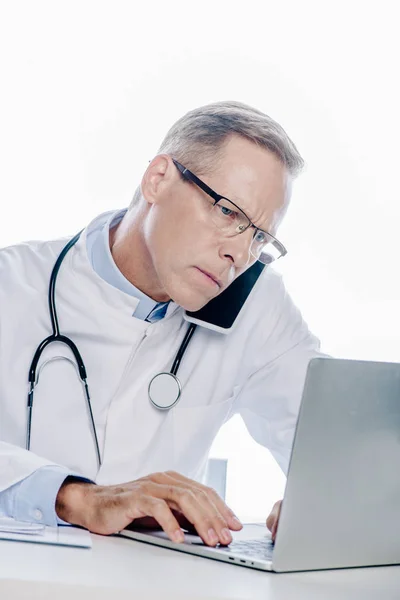 Médico Bonito Casaco Branco Falando Smartphone Usando Laptop Isolado Branco — Fotografia de Stock