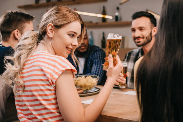 selective focus of smiling young woman holding glass of beer while sitting with multicultural friends in pub