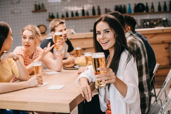 开朗的年轻女子拿着一杯点燃的啤酒 看着相机 而坐在酒吧的朋友附近 — 图库照片