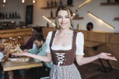 schöne Kellnerin in deutscher Tracht blickt in die Kamera und zeigt Willkommensgeste