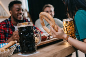 cropped view of woman holding pretzel while celebrating octoberfest with multicultural friends in pub