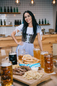 schöne junge Kellnerin in traditioneller deutscher Tracht hält die Hand auf der Hüfte und lächelt in die Kamera