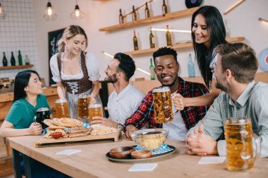 two attractive waitresses serving beer for multicultural friends in pub clipart