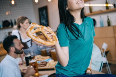 cropped view of smiling asian girl holding pretzel while celebrating octoberfest with multicultural friends clipart