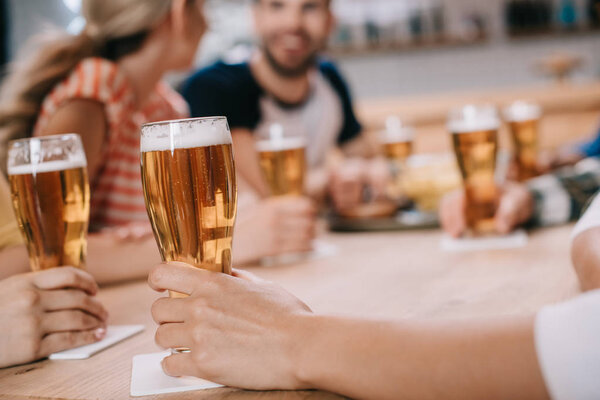 cropped view of women holding glasses of light beer while sitting together with friends in pub
