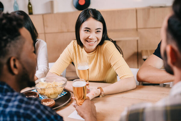 selective focus of smiling asian girl holding glass of light beer while sitting in pub near multicultural friends