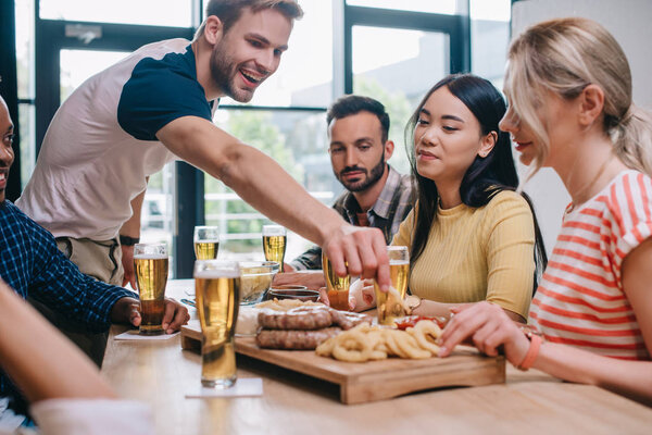 cheerful multicultural friends sitting at pub near tray with fried onion rings, sausages and french fries
