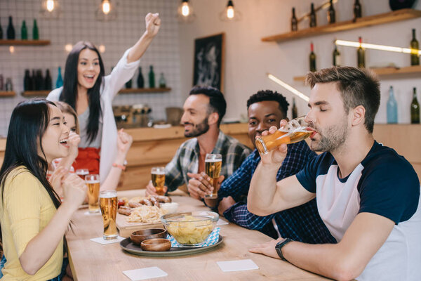 young man drinking beer with closed eyes while sitting in pub together with cheerful multicultural friends
