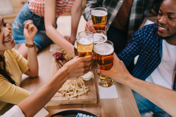 cropped view of multicultural friends clinking glasses of light beer in pub