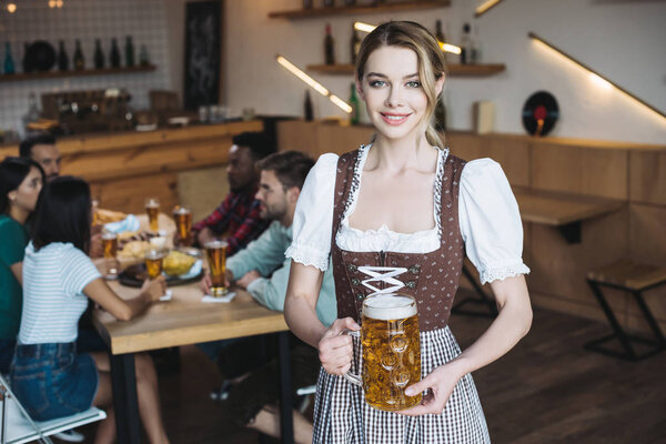 beautiful waitress in german national costume holding mug of light beer and smiling at camera
