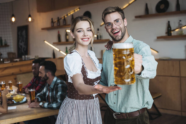 selective focus of handsome young man holding mug of light beer while standing near beautiful waitress in traditional german costume