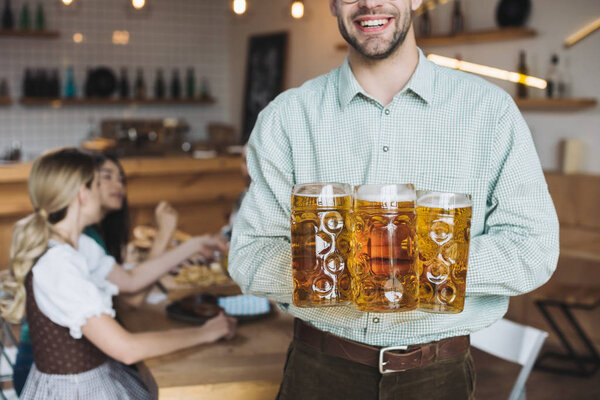 cropped view of young man holding mugs with beer and smiling at camera