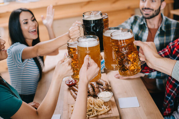 cheerful friends clinking mugs of beer while celebrating octoberfest in pub