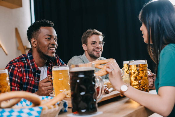 cheerful multicultural friends sitting near mugs with light and dark beer in pub