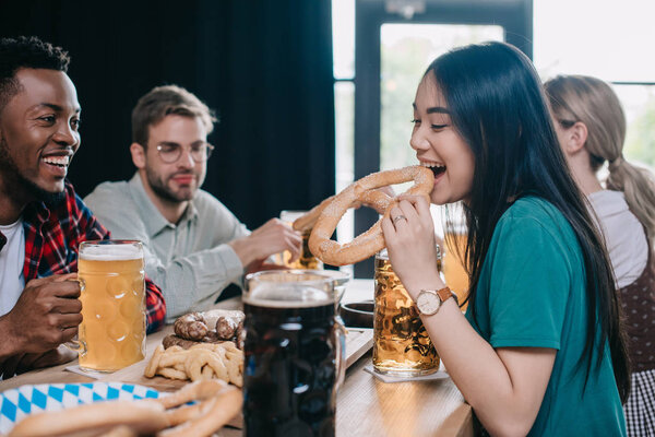 young asian woman eating pretzel while celebrating octoberfest with multicultural friends in pub