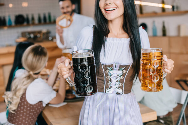 cropped view of waitress in traditional german costume holding mugs of light and dark beer