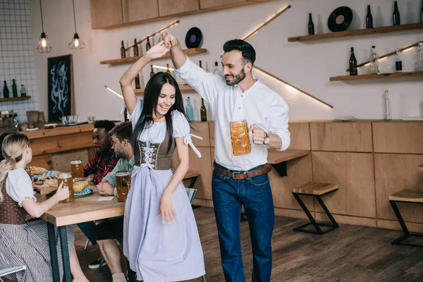 开朗的年轻人拿着一杯啤酒 同时跳舞与迷人的女人在传统的德国服装 — 图库照片