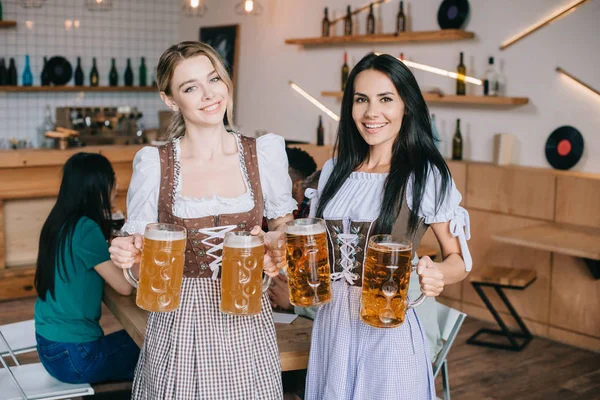 two beautiful waitresses in traditional german costumes holding mugs with beer and smiling at camera