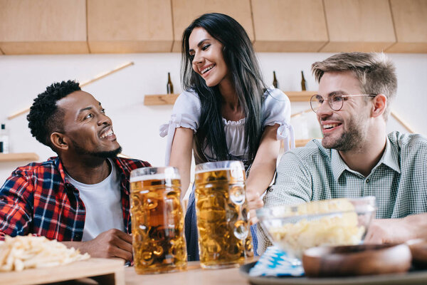 cheerful waitress in traditional german costume serving beer for multicultural friends in pub