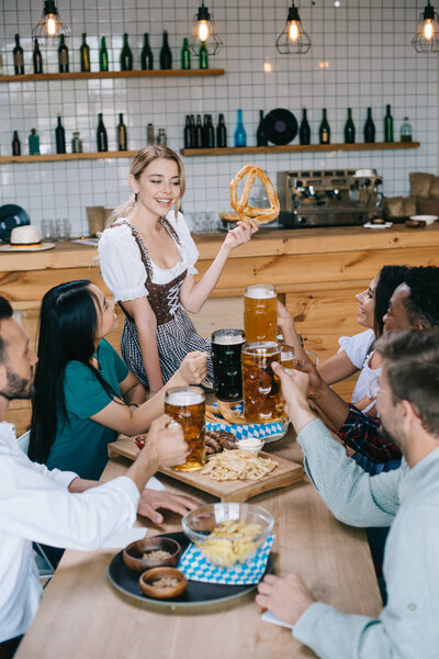 cheerful waitress in traditional german costume holding pretzel while standing near multicultural friends celebrating octoberfest in pub