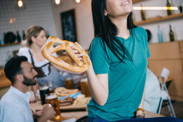 cropped view of smiling asian girl holding pretzel while celebrating octoberfest with multicultural friends