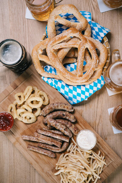 top view of fried sausages, onion rings, french fries, pretzels and mugs with beer on wooden table in pub