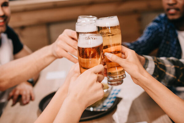 cropped view of multicultural friends clinking glasses with lager beer in pub