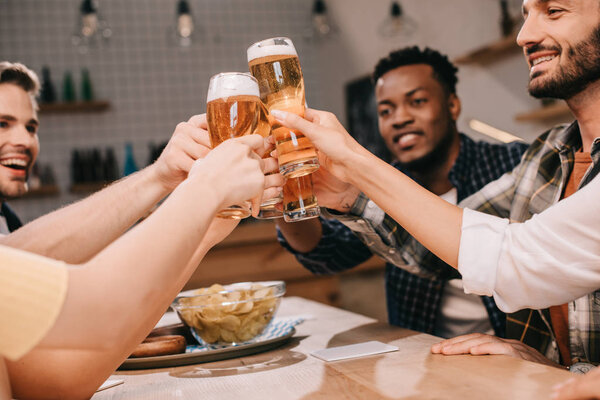 partial view of cheerful multicultural friends clinking glasses with lager beer in pub