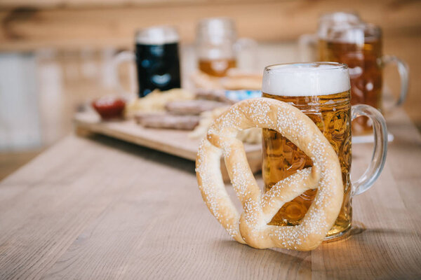 selective focus of mug with lager beer and pretzel on wooden table near tray with snacks in pub