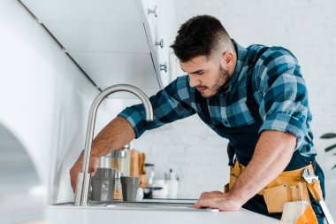 selective focus of handsome handyman working near sink in kitchen  clipart