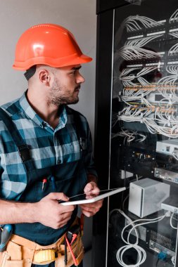 handsome bearded workman holding digital tablet and looking at wires and cables  clipart