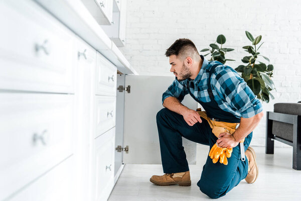 selective focus of handsome handyman with tool belt sitting near kitchen cabinet