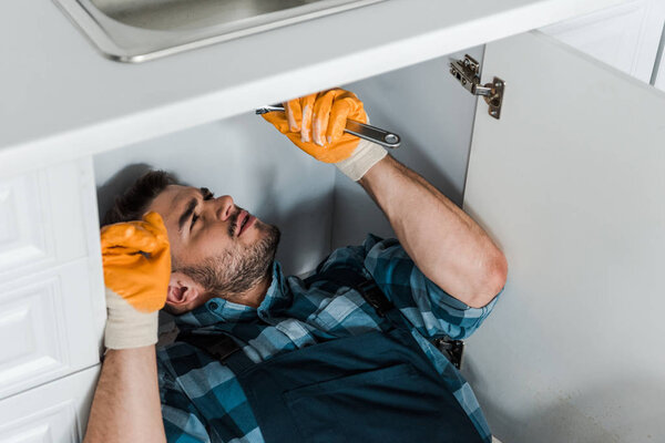 bearded repairman holding adjustable wrench while working in kitchen 