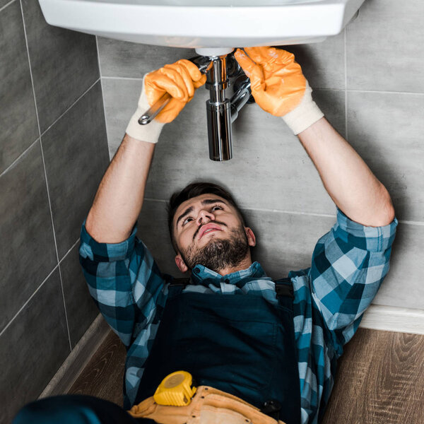 bearded repairman fixing water damage with adjustable wrench in bathroom 