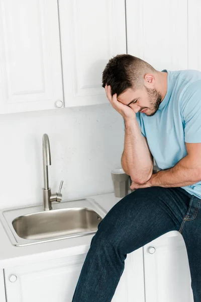 upset man touching face while looking at faucet in kitchen
