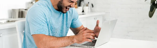 panoramic shot of angry man gesturing while sitting near laptop