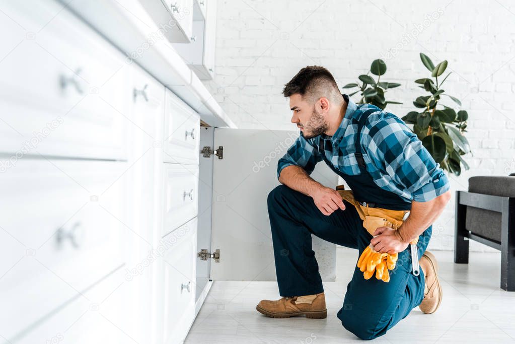 selective focus of handsome handyman with tool belt sitting near kitchen cabinet