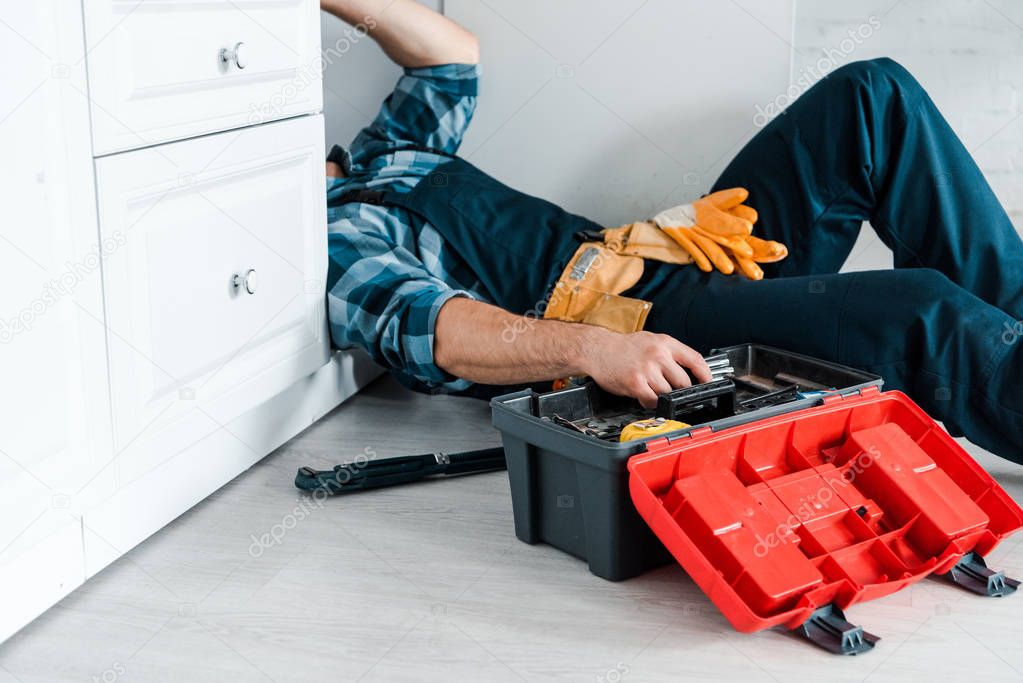 cropped view of repairman working in kitchen near toolbox while lying on floor 