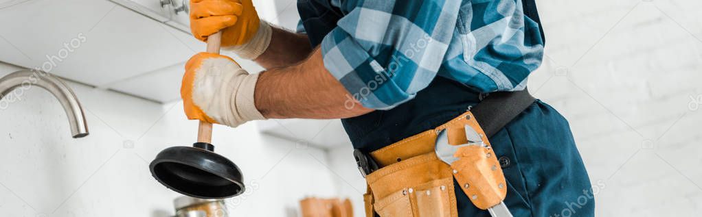 panoramic shot of handyman with tool belt holding plunger in kitchen 