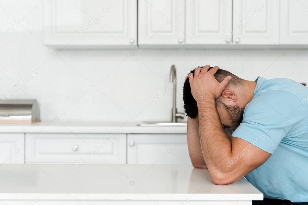 frustrated man touching head while sitting near table in kitchen 
