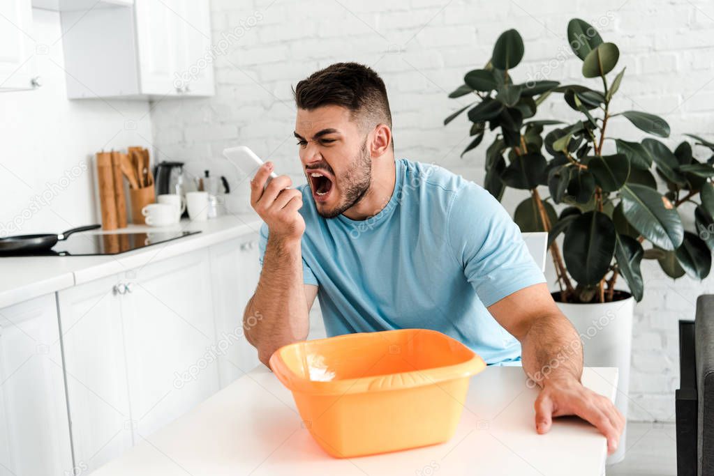 selective focus of upset bearded man screaming at smartphone near plastic wash bowl 