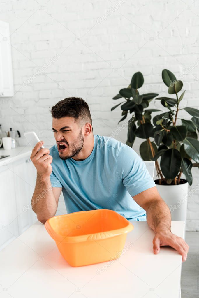 selective focus of upset bearded man screaming while holding smartphone near water pouring into plastic wash bowl 