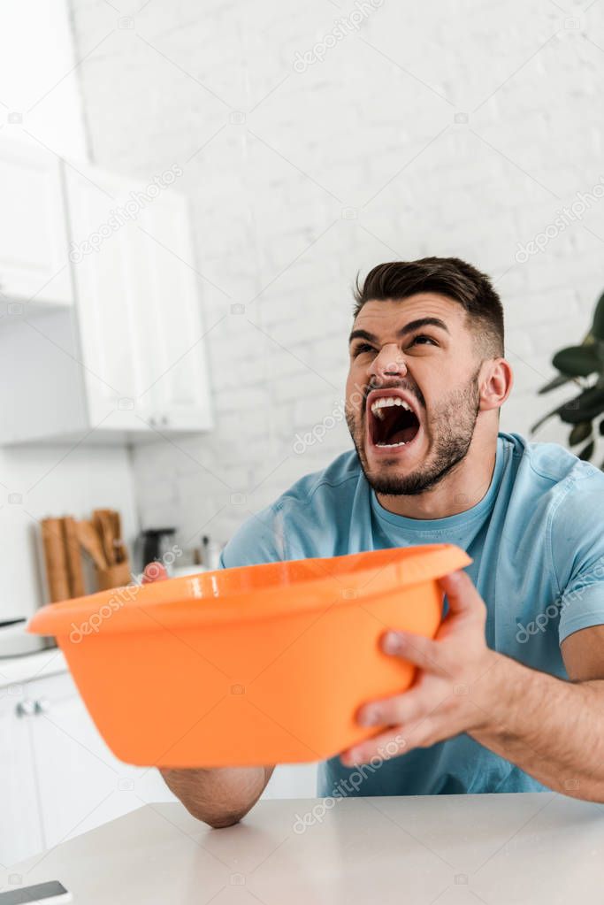 selective focus of bearded  man screaming while holding plastic wash bowl near pouring water 