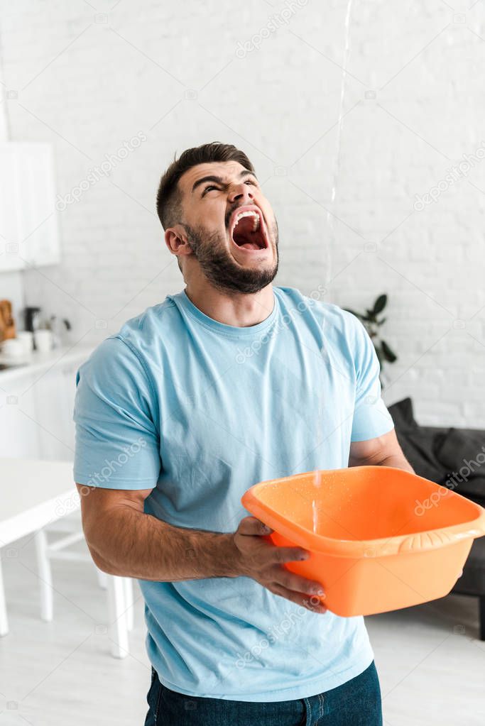 selective focus of upset man screaming while holding plastic wash bowl near pouring water 